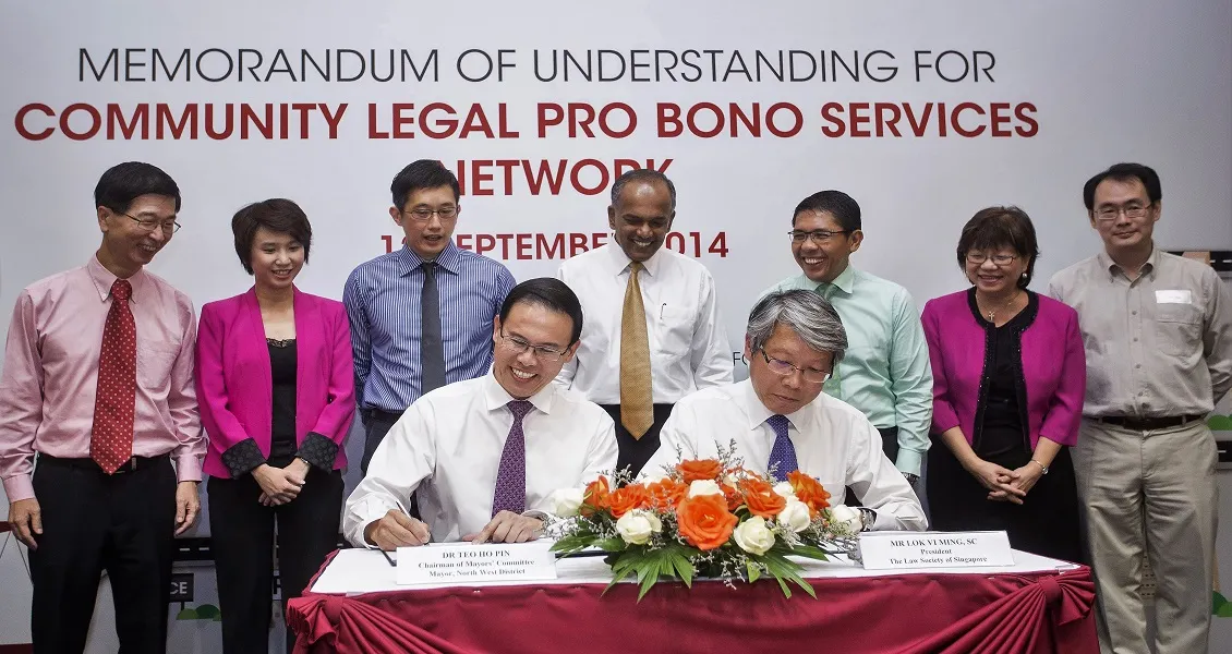 Several people stand behind two individuals signing documents at a table adorned with flowers; a banner is in the background.
